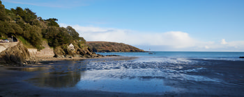 VIEW FROM BAY 3 The image shows a landscape photograph taken from Bay 3 overlooking a quiet coastal beach in Devon, England, United Kingdom. The view was captured in the late afternoon during the winter season, as indicated by the low angle of the sunlight and the bare vegetation on the distant hillside. The beach is bordered by rocky cliffs and a stretch of sand with shallow pools of water reflecting the sky. The coastline, lined with trees and a stone wall, curves gently towards a headland projecting into the sea. The image highlights the natural beauty of the Devon coast, with clear skies and calm waters visible in the distance.